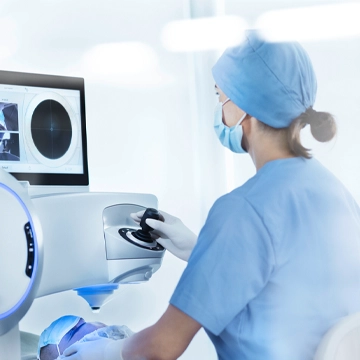 The image depicts what appears to be a healthcare professional, likely a woman based on her clothing and posture, sitting at a computer workstation. She is wearing medical scrubs and focusing intently on the screen. In front of her are various bits of medical equipment including an ultrasound machine with its screen displaying images or medical data. The setting suggests that she is in a medical facility like a hospital or doctor's office, given the professional attire and the presence of medical devices. It is likely that she is studying medical images or analyzing patient data as part of her job.