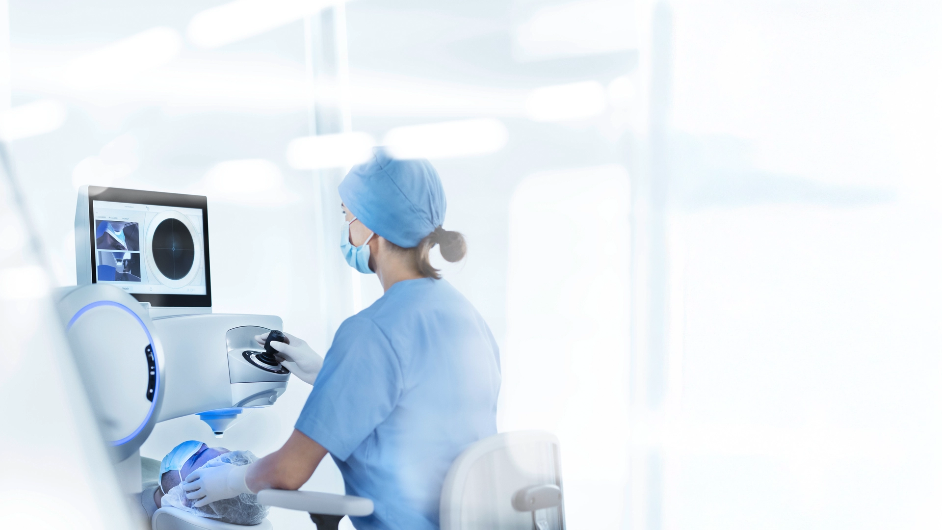 The image depicts what appears to be a healthcare professional, likely a woman based on her clothing and posture, sitting at a computer workstation. She is wearing medical scrubs and focusing intently on the screen. In front of her are various bits of medical equipment including an ultrasound machine with its screen displaying images or medical data. The setting suggests that she is in a medical facility like a hospital or doctor's office, given the professional attire and the presence of medical devices. It is likely that she is studying medical images or analyzing patient data as part of her job.